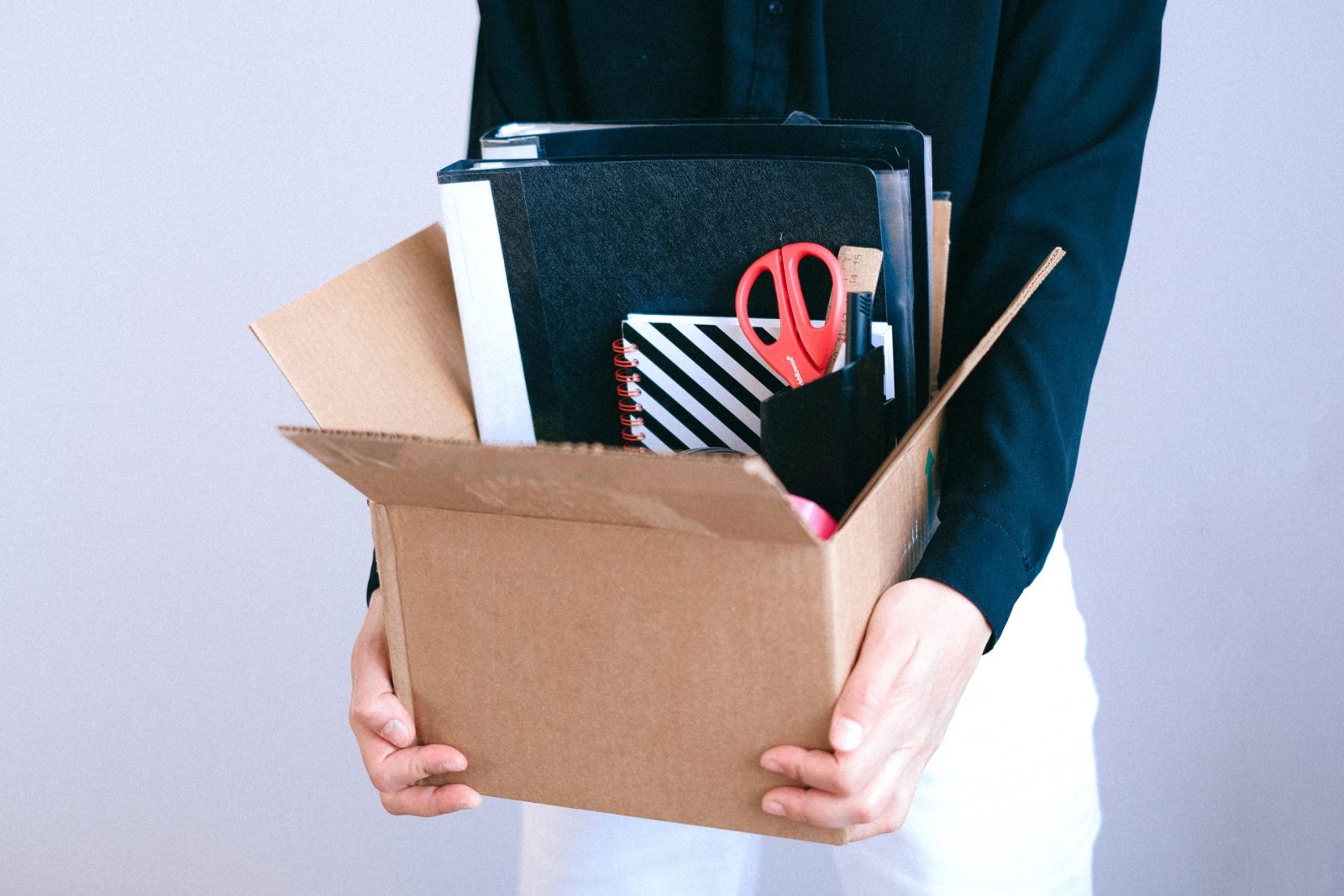 Persona sujetando una caja de cartón con carpetas, libretas y material de oficina al dejar su puesto de trabajo